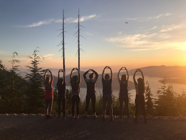 An aerial view of a group of Americans performing a yoga class outdoors during sunset, with a natural background featuring scenic mountains in the distance.