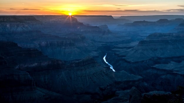A dramatic view of the Black Canyon of the Gunnison from a high vantage point. The sun is setting, casting long shadows across the canyon walls and highlighting the intricate patterns of the rock formations. The Gunnison River can be seen flowing far below.