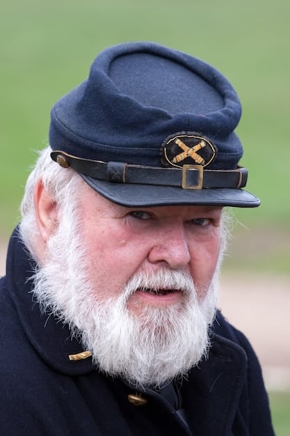 Close-up of a reenactor dressed as a Continental Army soldier. He's adjusting his tricorn hat and holding a flintlock musket. The background shows a tent camp with other reenactors preparing for a demonstration.