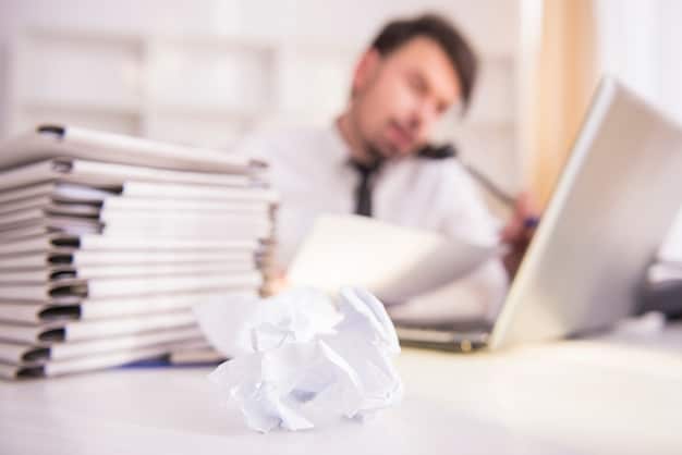 A cluttered office desk overflowing with paperwork and documents, with a stressed-looking business person in the background blurred. This symbolizes the burden of regulatory compliance.